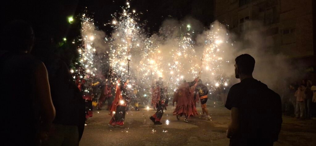 Participants dressed as devils holding spinning pitchforks that shower bright white and orange sparks over a crowded street during a traditional Catalan correfoc festival at night.